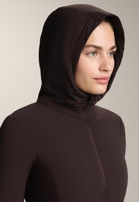 Huvjacka i mörkbrun, tillverkad av slät, lättviktsmaterial. Har en hel dragkedja fram och en figurnära design.