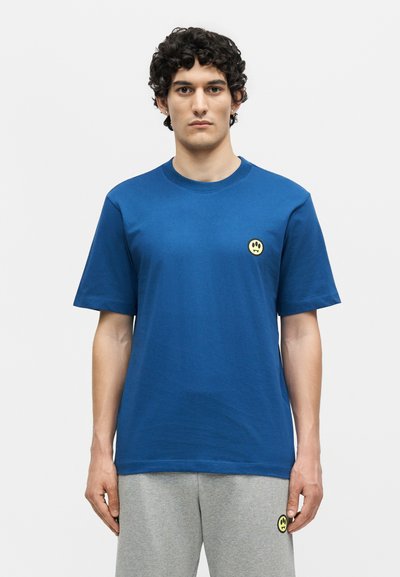 Νέος άνδρας με σγουρά μαλλιά που φοράει μπλε μπλουζάκι και γκρι παντελόνι φόρμας, και τα δύο με μικρό κίτρινο λογότυπο με χαμογελαστό πρόσωπο.