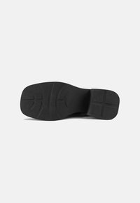 Semelle de chaussure noire à bout et talon carrés, avec des lignes de grip courbées à l’avant-pied et un motif en croix sur le talon, sur fond uni.