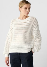 Weißer Strickpullover mit weiten, offenen Maschen und gerippten Bündchen. Leicht oversized Schnitt mit Rundhalsausschnitt. Getragen mit dunkler Hose.