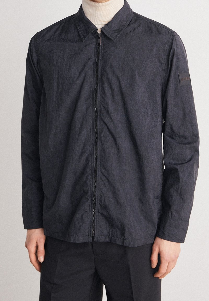 Homme portant une veste zippée bleu marine foncé par-dessus un col roulé beige, associée à un pantalon noir sur un fond clair uni.