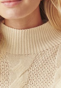 Krēmkrāsas adīts džemperis ar masīvu ribotu apkakli, detalizētām kabelēm un teksturētu auduma struktūru vieglā materiālā.