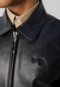 Sort læderjakke med spids krave, frontlynlås og præget "Schott NYC" logo, båret over en beige skjortekrave.