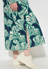 Blommig midiklänning med grönt och krämfärgat lövmönster på en marinblå bakgrund, med en slits på sidan, kombinerad med krämfärgade sneakers.