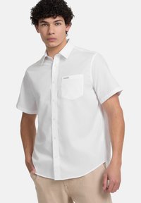 Korte mouwen witte blouse gemaakt van soepel stof, met een borstzak en een label boven de zak, en een afgeronde zoom.