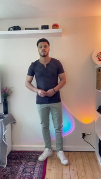Camisa henley de manga corta a rayas en tonos azul marino, pantalones verdes claros y zapatillas blancas. De pie sobre un suelo de madera con una alfombra estampada y estanterías.