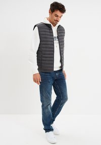 Chaleco de plumas gris con cuello alzado, cierre frontal y costuras horizontales, llevado sobre una sudadera blanca y jeans azules. Zapatillas blancas.