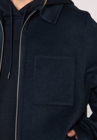 Persona che indossa un cappotto zip-up di un blu scuro sopra un hoodie blu, con una grande tasca quadrata sul petto del cappotto e una mano infilata nella tasca del cappotto.