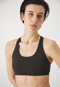 Svart sports-bh med scoop-halsutskjæring og brede stropper, laget av glatt, elastisk materiale. Har en tettsittende design og minimal sømmer.