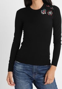 Schwarzer Pullover mit langen Ärmeln aus Strickstoff, der florale Verzierungen in Rot, Weiß und Grün auf der linken Schulter aufweist.