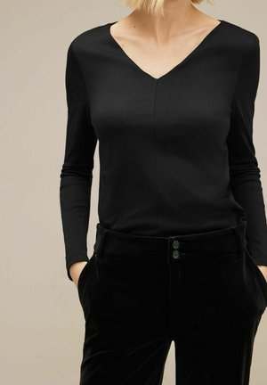 Vrouw draagt een zwart langarmig V-hals shirt en zwarte broek met handen in de zakken tegen een beige achtergrond.