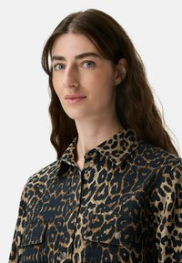 Frau mit langen braunen Haaren, die ein braunes und schwarzes Leopardenmuster-Hemd mit Knöpfen und Fronttaschen trägt, vor einem schlichten Hintergrund.