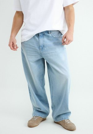 Homme portant un jean large bleu clair, un t-shirt blanc et des baskets beiges, debout devant un fond uni.