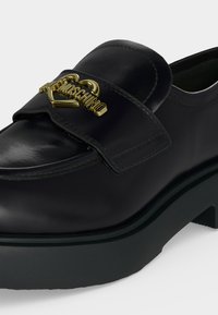 Μαύρο δερμάτινο loafer με χοντρή σόλα και χρυσή λεπτομέρεια σε σχήμα καρδιάς που φέρει το όνομα της μάρκας Moschino στο πάνω λουράκι.
