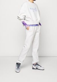 Vitt fleece-tröja med blå text "Var snäll", lager på lager med en lila tie-dye långärmad tröja. Kombinerad med vita sweatpants och grå sneakers.