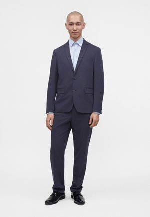 Homme debout portant un costume bleu marine foncé avec une chemise bleu clair et des chaussures habillées noires sur fond blanc.
