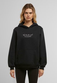 Felpa nera con cappuccio, realizzata in tessuto morbido, dotata di una tasca frontale e testo bianco in arabo e inglese che recita "Ama te stesso prima di tutto."