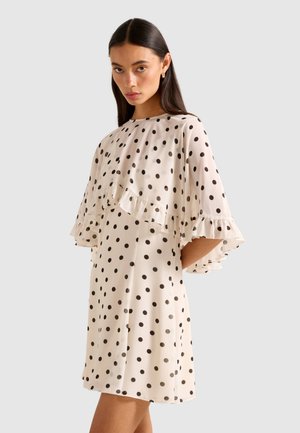 Woman wearing a white dress with black polka dots and ruffled cape sleeves, standing against a plain light gray background.