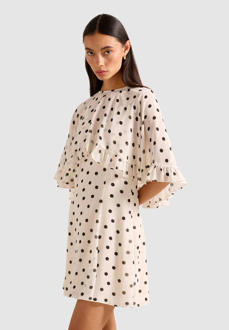 Woman wearing a white dress with black polka dots and ruffled cape sleeves, standing against a plain light gray background.