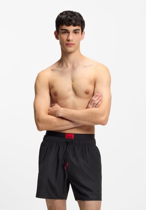 Młody mężczyzna z krótkimi ciemnymi włosami stoi topless w czarnych kąpielówkach z czerwonym logo Hugo, ze skrzyżowanymi ramionami, na neutralnym białym tle.