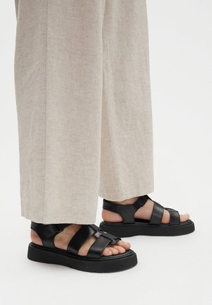 Schwarze Leder-Sandalen mit mehreren Riemen und flachen Sohlen, mit einer Schnallendetail. Getragen zu hellfarbenen, weit geschnittenen Hosen.