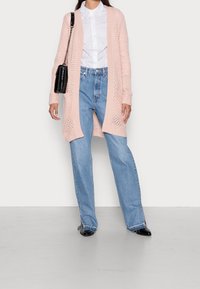 Mujer vestida con un cardigan largo de punto rosa, camisa blanca de botones, jeans de color azul claro, zapatos negros de punta y llevando un bolso negro de hombro.