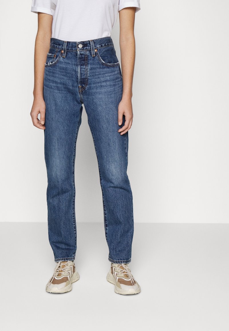 Personne portant un jean taille haute bleu et des baskets beiges épaisses, debout devant un fond blanc uni.
