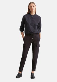 Zwarte blouse met stippen, ronde kraag en knoopsluiting. Zwarte cargobroek met drawstring taille en twee zijzakken. Zwarte schoenen.