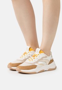 Osoba obutá v béžových a hnědých teniskách s bílou podrážkou a oranžovými akcenty, stojící s nohama blízko u sebe na bílém pozadí.