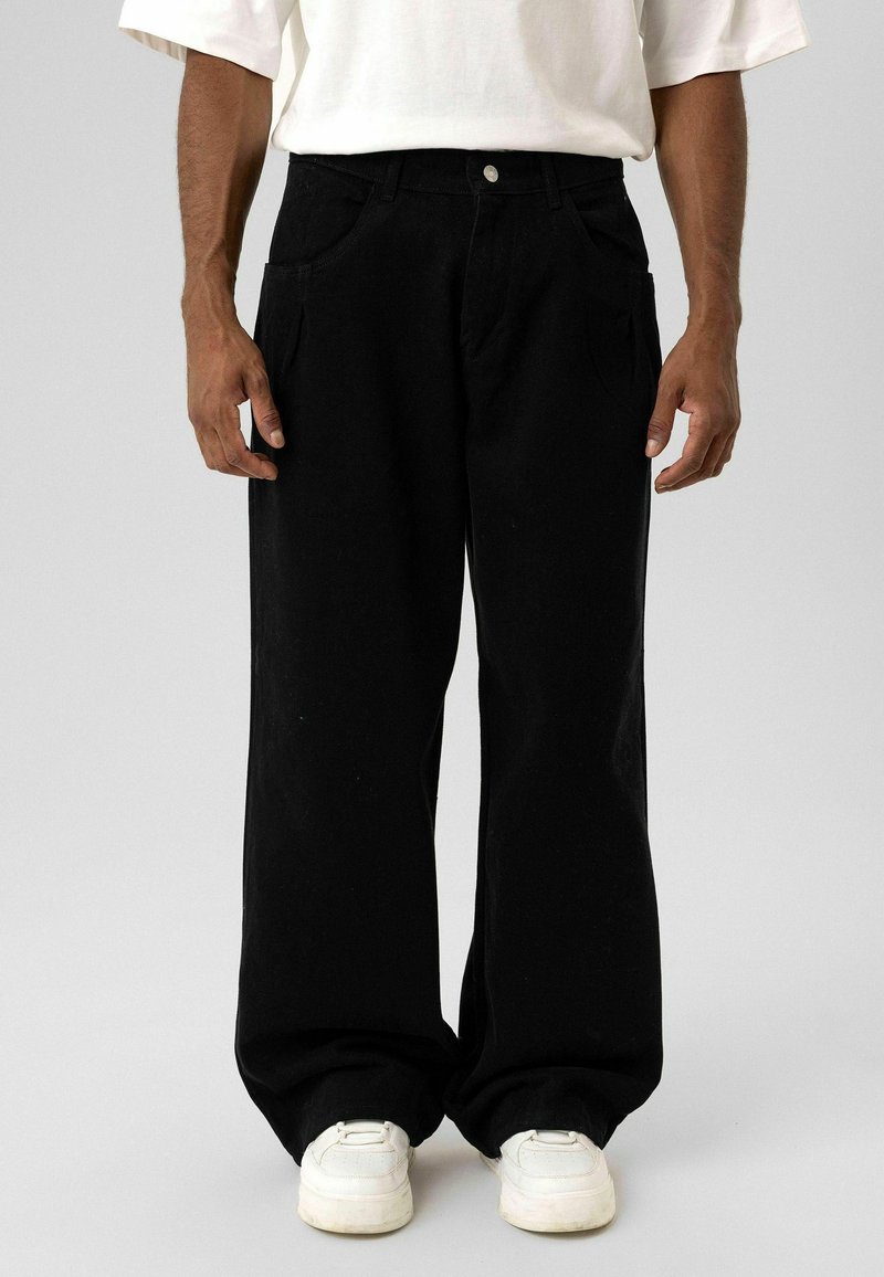 Zwarte wijd uitlopende broek van denim, met twee steekzakken aan de voorkant en een knoopsluiting, gecombineerd met witte sneakers.