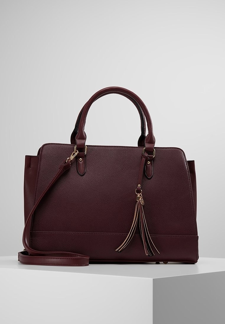 Sac à main en cuir bordeaux foncé avec double poignée, bandoulière amovible et pompon décoratif, posé sur un socle blanc sur fond gris.
