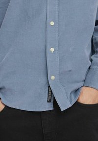 Hellblaues Cordhemd mit Knopfleiste, ausgestattet mit weißen Knöpfen und einem seitlichen Etikett, kombiniert mit schwarzen Jeans. Strukturierter Stoff.