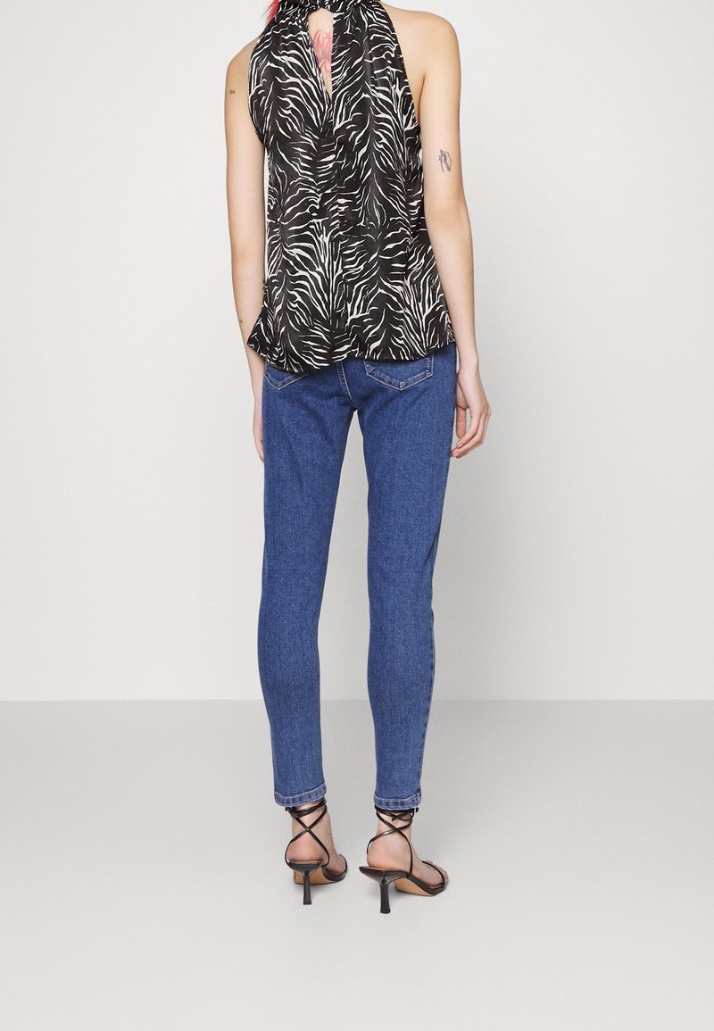 Femme portant un haut sans manches à imprimé zèbre noir et blanc, un jean skinny bleu et des sandales à talons hauts noires à lanières, se tenant de dos par rapport à la caméra.