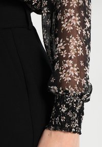 Blouse noire à fleurs présentant une manche transparente et aérienne ornée de petites fleurs blanches et roses, avec un poignet noir froncé.