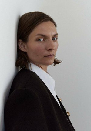 A person with medium-length brown hair wears a dark coat with gold buttons and a white shirt, leaning against a light wall.