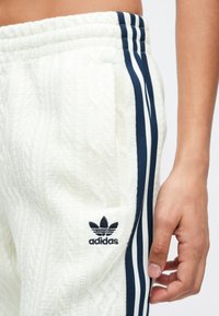 Close-up de uma pessoa a usar calças brancas da Adidas com riscas e logótipo em azul-marinho, com a mão descansando perto do bolso do lado direito.