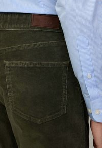 Pantaloni in velluto a coste verde oliva con una tasca posteriore e etichetta in pelle. Abbinati a una camicia a quadri azzurri chiari, con polsini abbottonati.