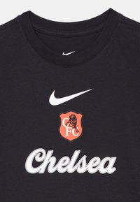 Μαύρη βαμβακερή μπλούζα με λευκό λογότυπο Nike, κόκκινο και μαύρο σήμα της Chelsea FC και έντονο λευκό κείμενο "Chelsea". Κλασικός σχεδιασμός με στρογγυλή λαιμόκοψη.