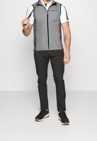 Grau gefütterte Golfweste mit durchgehendem Reißverschluss, seitlichen Einsätzen und Logo auf der Brust. Getragen über einem weißen Polo und kombiniert mit schwarzen Hosen und Sneakers.