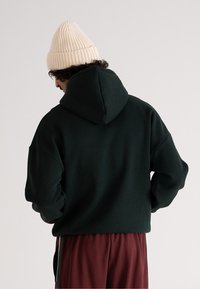 Felpa verde scuro con un ampio cappuccio, texture morbida, vestibilità rilassata, indossata con pantaloni borgogna e un berretto a coste beige in testa.