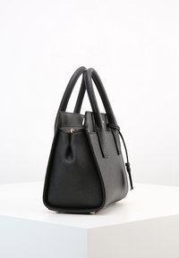 Borsa a tracolla in pelle nera strutturata con manici doppi, zip dorata e piccoli piedini in metallo, posizionata su un piedistallo bianco contro uno sfondo semplice.