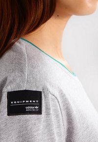 Person som bär en grå Adidas Equipment-tröja med en svart etikettpatch på övre ärm med vit text.