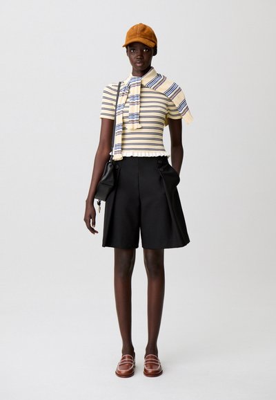 Top de punto amarillo a rayas con bajo fruncido y una bufanda, combinado con pantalones cortos negros plisados. Completa el conjunto una gorra color canela y mocasines marrones.