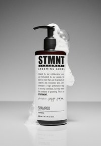 Svartvit schampoflaska med pumpdispenser. Etiketten har fet text med varumärkesnamn och instruktioner. Bubblor nära toppen.