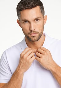 Valkoinen polo-paita, jossa on klassinen kaulus, malli säätää kaulusta käsillään. Sujuva kangas, jossa on hienovarainen rakenne.