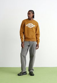 Brauner Sweatshirt mit weißem "Dickies"-Logo, grauen Cordhosen und schwarzen Turnschuhen, ausgestattet mit einem lockeren Schnitt und strukturierten Materialien.