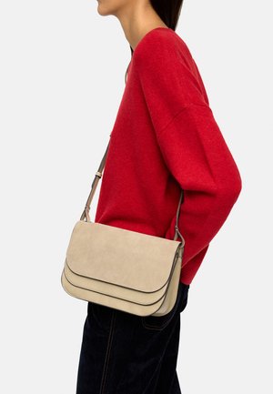 Kobieta w czerwonym swetrze i czarnych spodniach nosi na ramieniu beżową torbę na ramię z klapką i regulowanym paskiem.