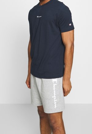 Mężczyzna w granatowej koszulce Champion i jasnoszarych szortach Champion stoi na jednolitym tle.