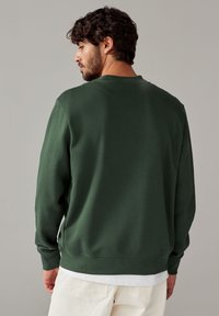 Dunkelgrüner Sweatshirt aus weicher Baumwolle, mit Rundhalsausschnitt und gerippten Bündchen. Das Model steht mit dem Rücken zur Kamera und trägt cremefarbene Hosen.