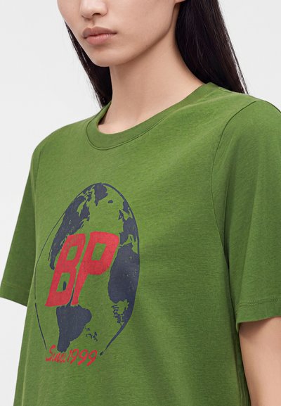 Γυναίκα που φοράει πράσινο T-shirt με μπλε και κόκκινη γραφική απεικόνιση γης και κόκκινο κείμενο "BP" και "Sun 1999" στο στήθος.
